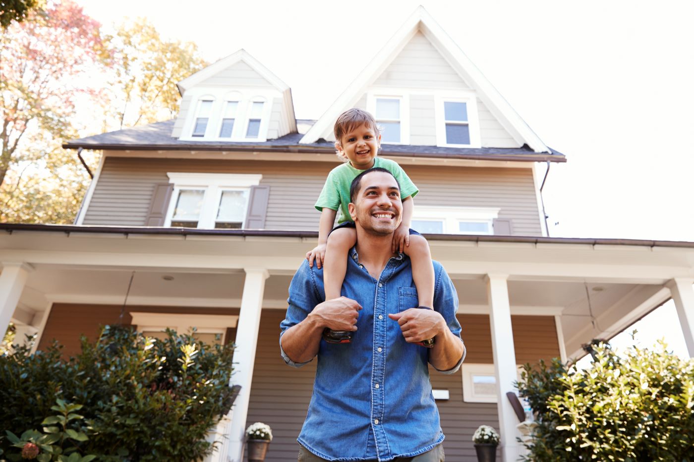 A man with his young son sitting on his shoulders Infront of his house.